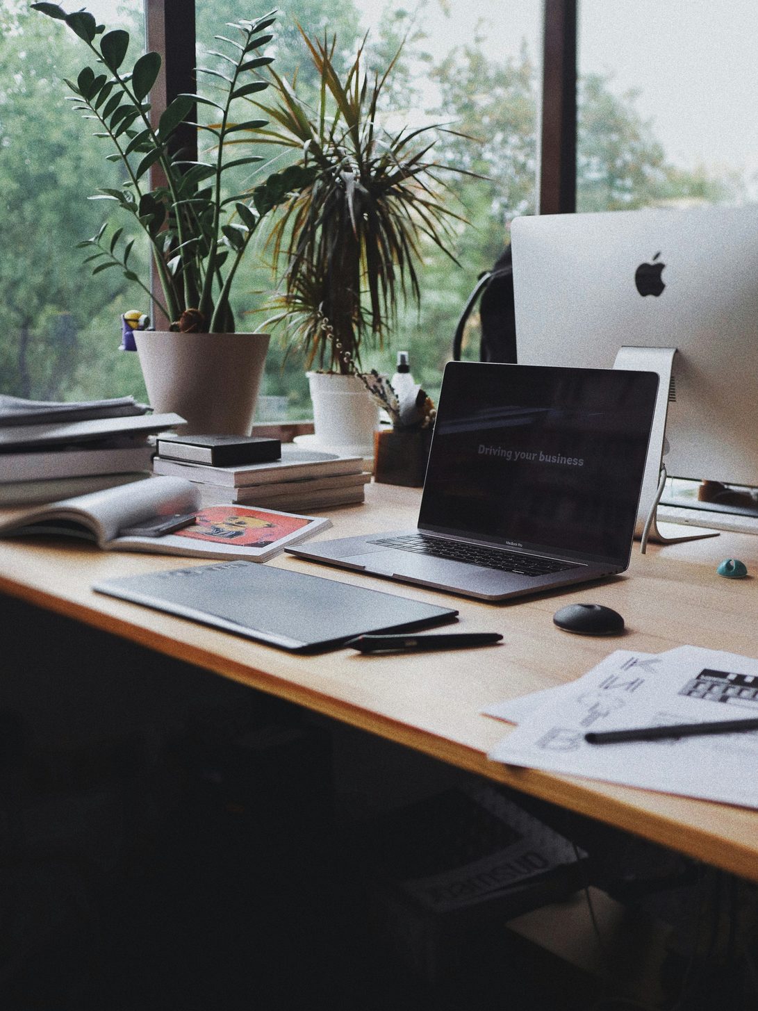 A wooden desk beside a window with an open laptop, a monitor, stacked books and sketch papers