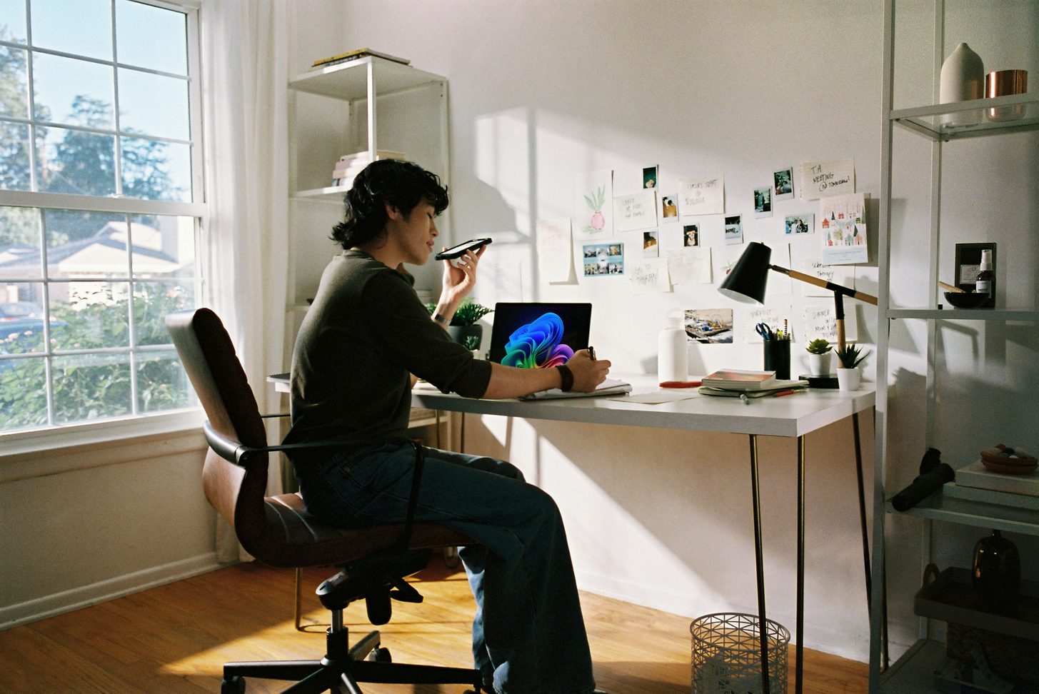 A person at a bright desk speaking on the phone while taking notes beside an open laptop