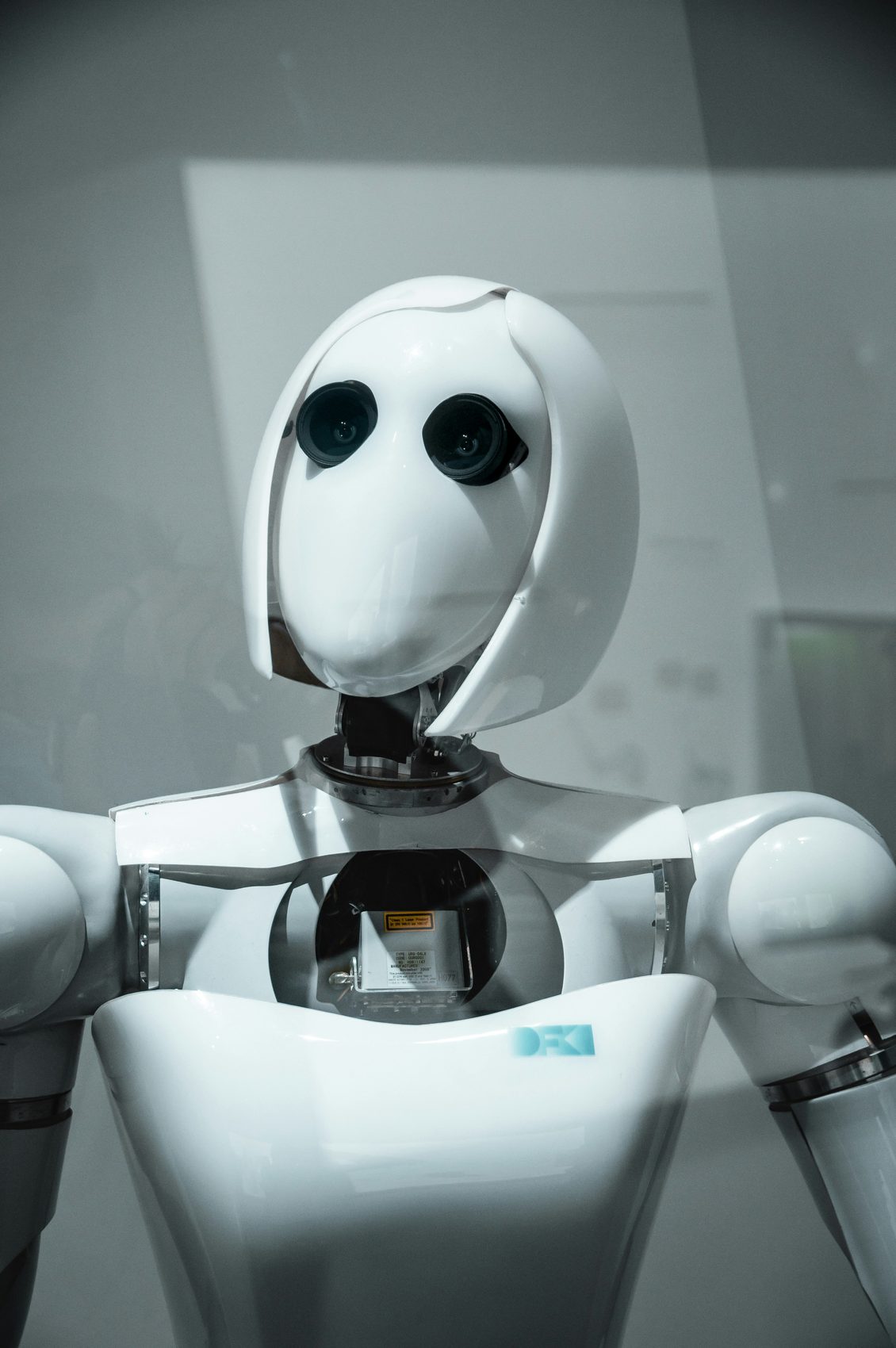 A white humanoid robot with dark lens eyes photographed head-on against a pale grey backdrop