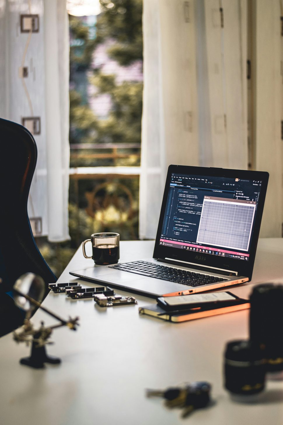 An open laptop showing a code editor on a desk with a coffee mug, circuit boards and notebooks