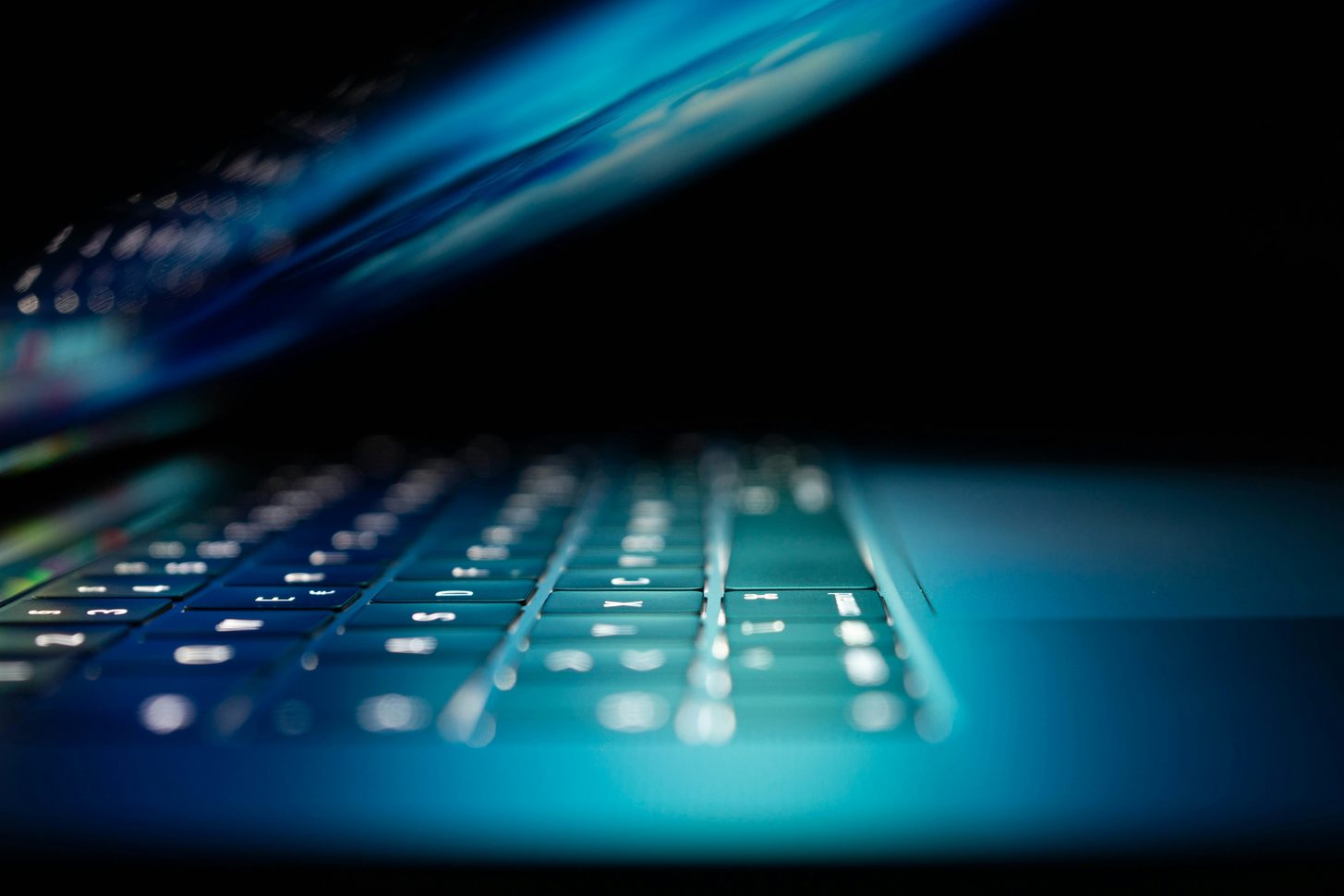 Extreme close-up of a backlit laptop keyboard glowing turquoise in a darkened room
